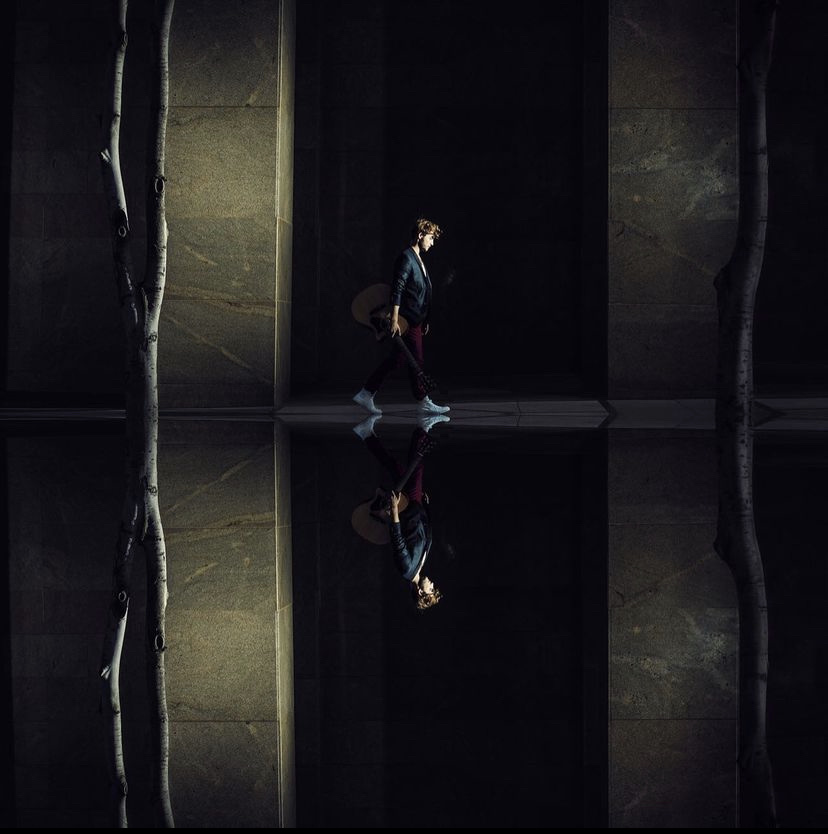 A musician carrying a guitar through a shaft of light, mirrored in reflection